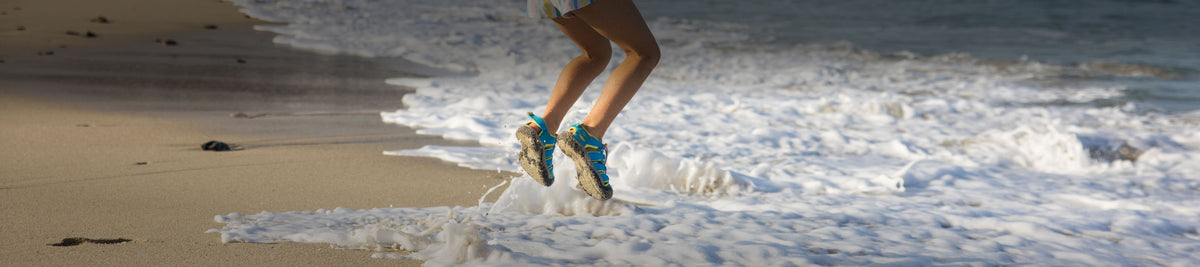 Two feet wearing KEEN kids' water shoes, shown jumping by the water on a beach shore.