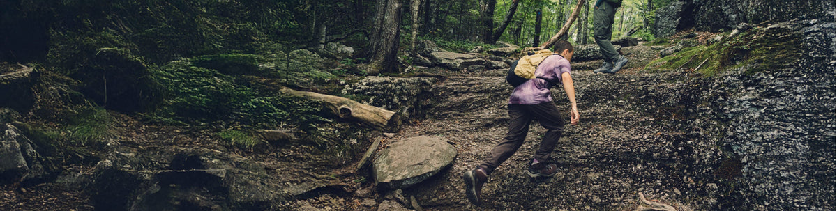 A hiker's feet shown wearing beige hiking boots, walking across rocky and grassy terrain.