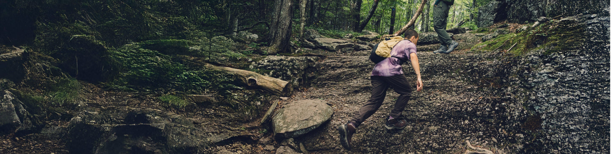 A hiker's feet shown wearing beige hiking boots, walking across rocky and grassy terrain.