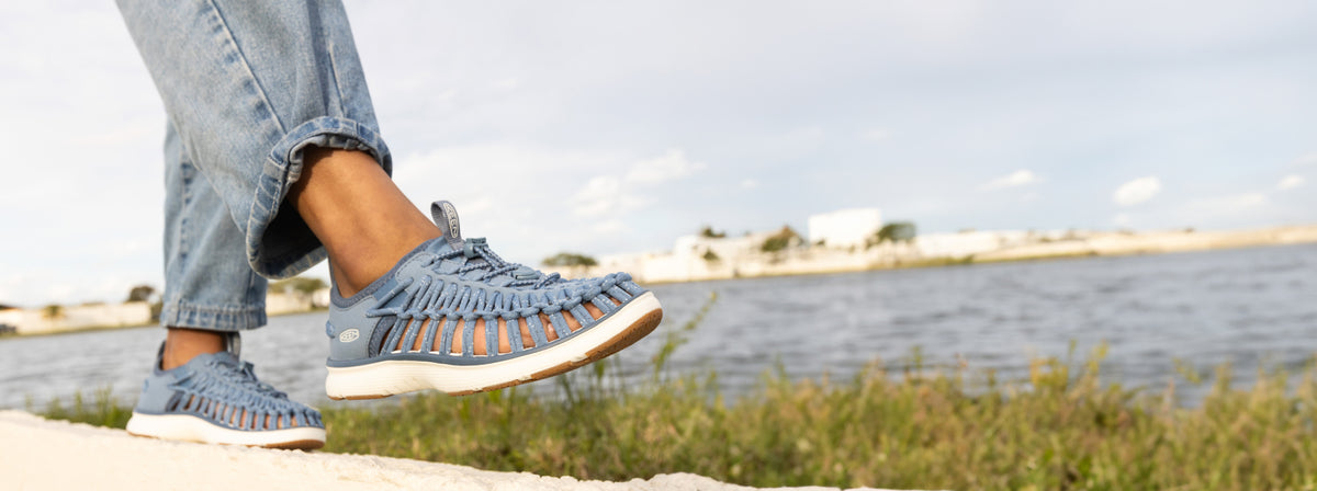 Knee down shot of woman wearing light blue Uneek O3 sandals and jeans walking on coastal boardwalk.