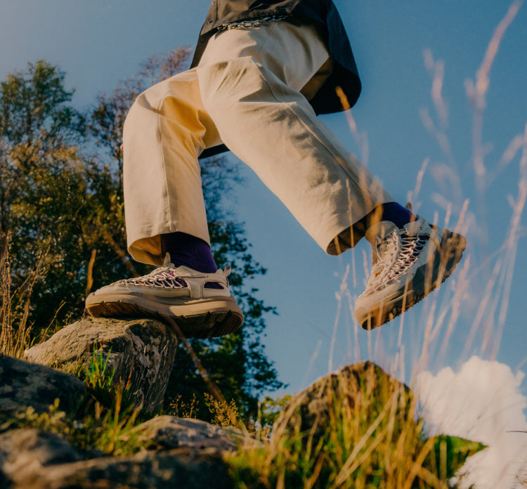 Low-angle shot of a person in tan pants and purple socks walking over rocks in woven KEEN sneakers against a blue sky.