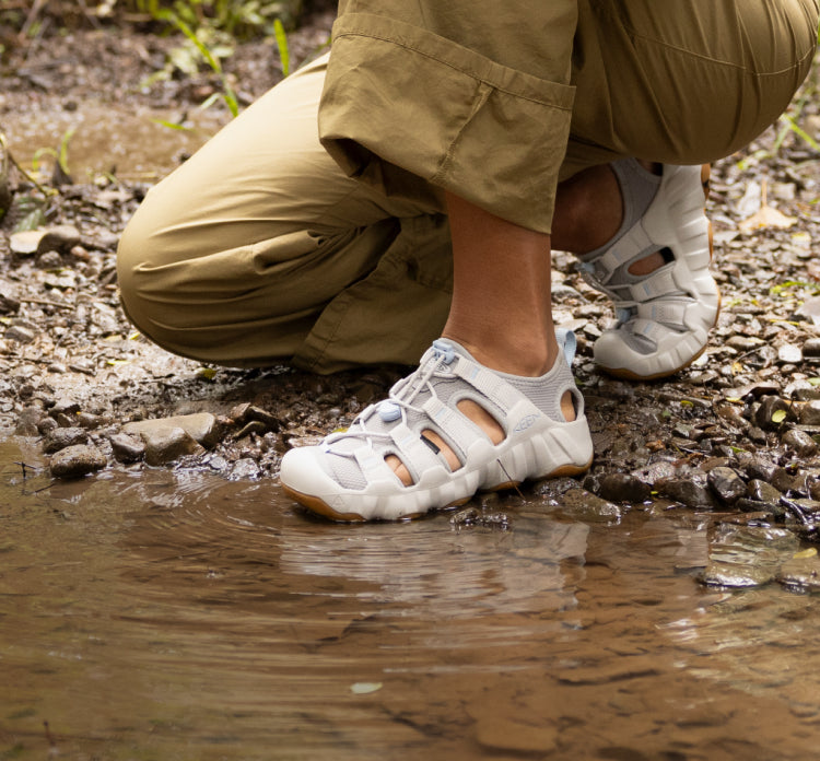 A person kneeling by the edge of a lake, wearing white Hyperport H2 sandals.