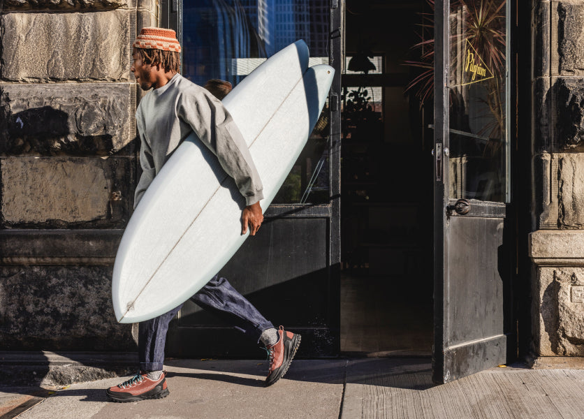 A person carrying a surfboard out of a shop, wearing KEEN sneakers.