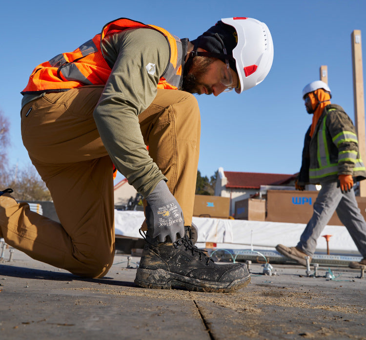 A man on a job site, kneeling down and securing the laces on his work boot.