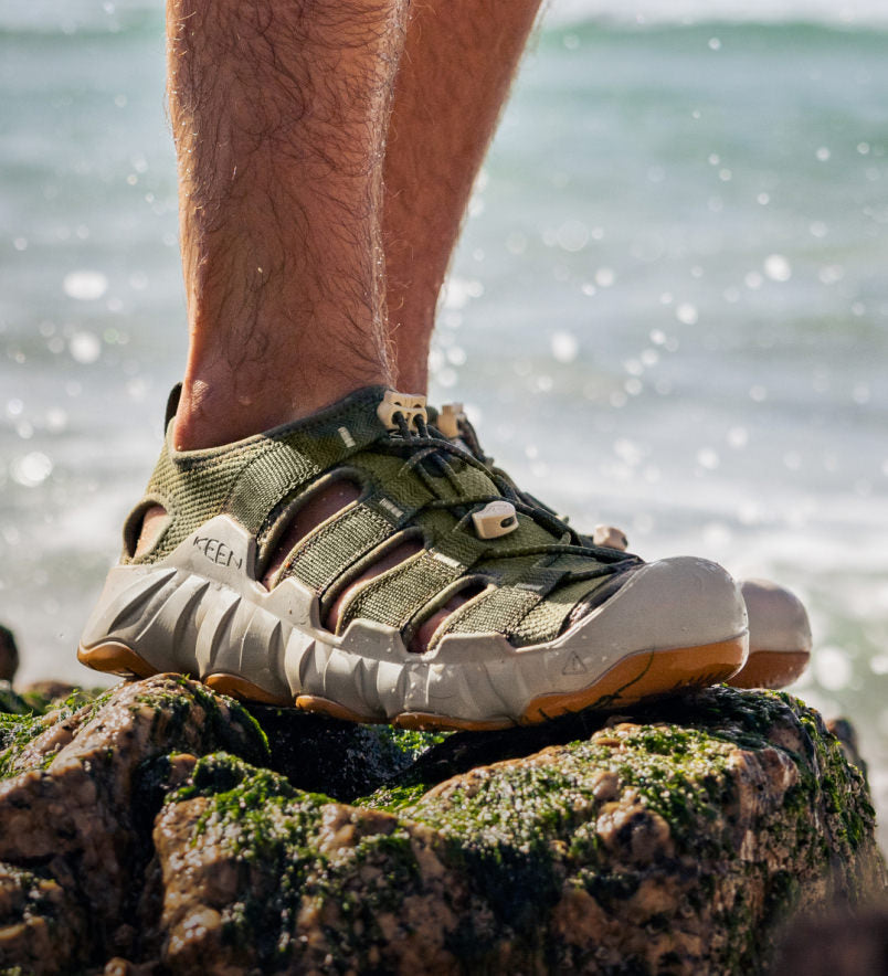 Knee-down shot of man wearing green and grey Hyperport water sandals and standing on mossy rock near ocean 