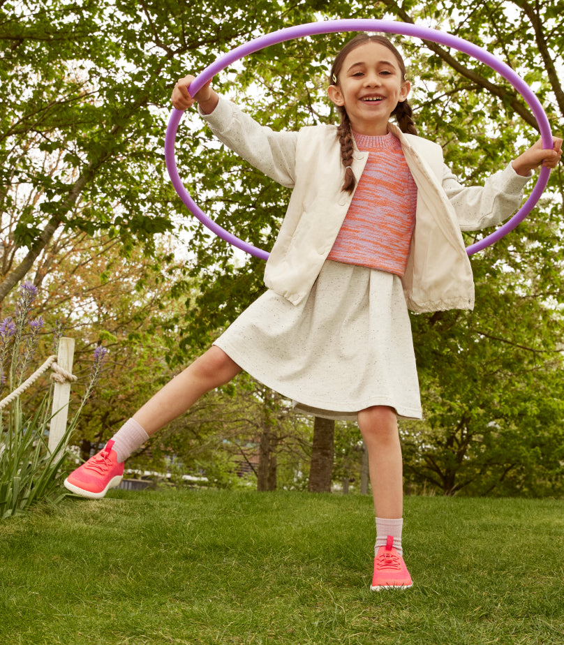 Lifestyle shot of little girl in white skirt and jacket with a pink sweater playing with a purple hula hoop while wearing pink knx knit sneakers in grassy park. 
