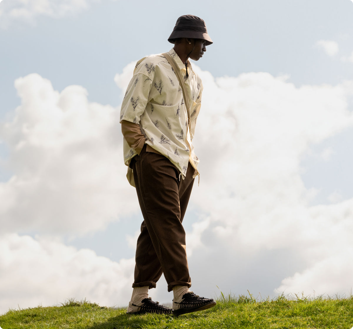 Man wearing brown pants and brown Uneek sandals walking across a grassy field with big clouds behind him. 