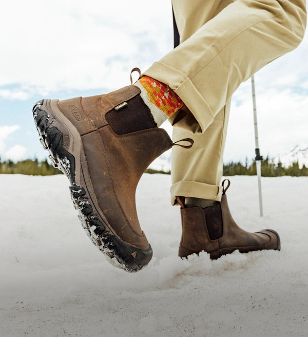 A person wearing a pair of brown KEEN boots, walking through snow.
