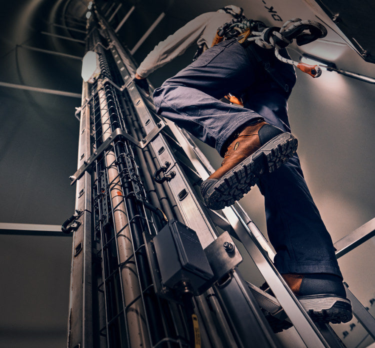 A worker climbing a tall, metal ladder, wearing a utility belt and a pair of KEEN Camden boots.