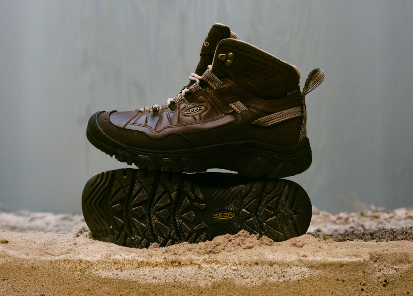 A pair of brown KEEN hiking boots atop a sandy surface.